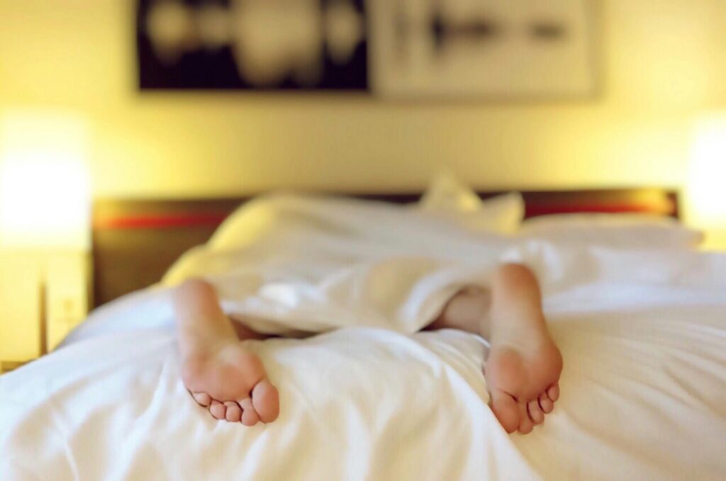 A person relaxing on a bed with feet under white sheets, conveying serenity. Yu Sleep Supplement